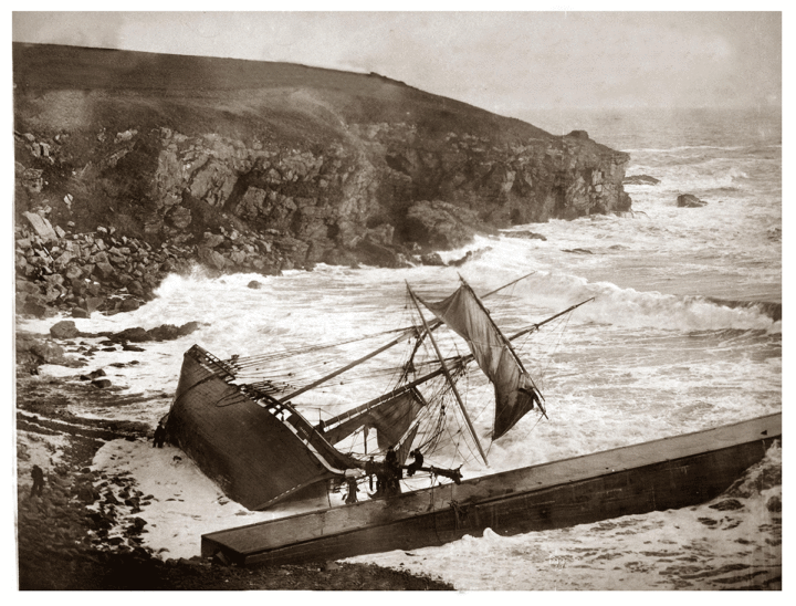 Sepia toned photograph of a shipwreck on its side lying on a beach next to rocky cliffs.