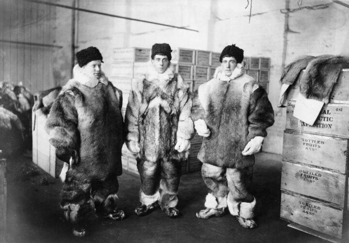 Monochrome photograph of three people in thick fur coats standing amongst stacked boxes in a store room.