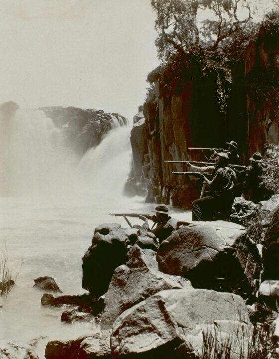 Sepia toned photograph of a group of snipers in military clothing standing on some rocks next to a waterfall. They are pointing their guns towards our left.
