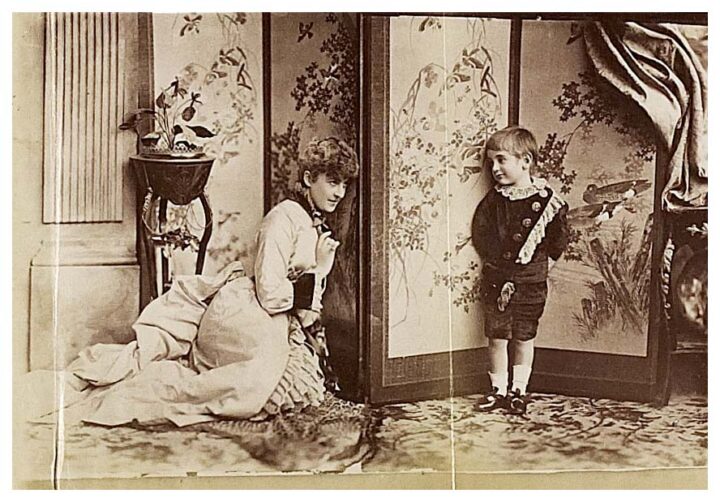 Sepia toned photograph of a woman kneeling on a carpeted floor next to a young boy in an opulently decorated room. They are looking at each other rather than at the camera.