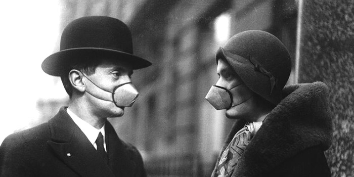 Two people stand opposite each other wearing gas masks. The photograph is from the 1920s.