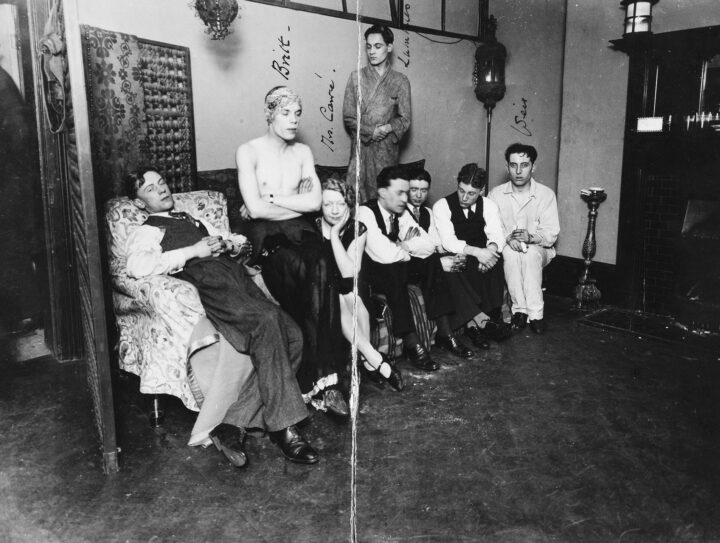 Monochrome photograph of a group of people sitting on a sofa in a living room. Most of them look away from the camera. A man labelled 'Britt' in pen sits shirtless with his arms crossed, wearing a long skirt and a headscarf.