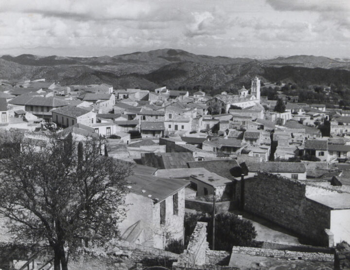 Monochrome photo of view from hill overlooking a Cypriot village with flat roofed houses, a church and the rolling hills beyond. 
