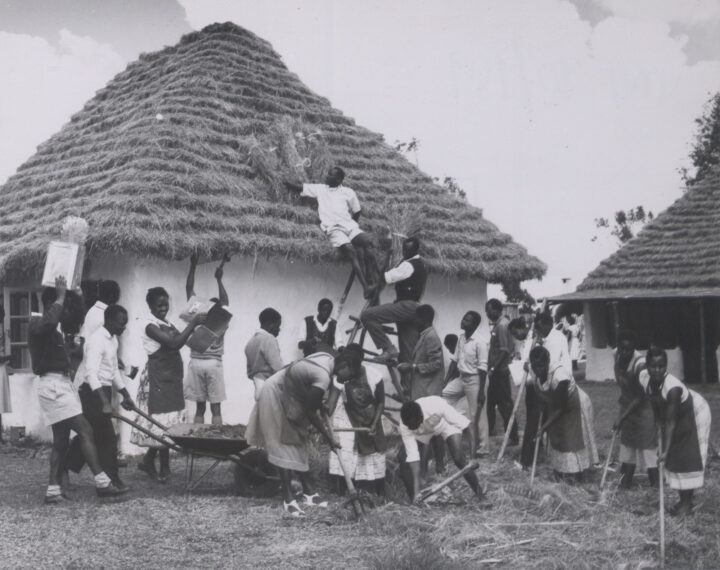 Black and white photograph of young men, women and children working on the cone-shaped thatch of a building.
