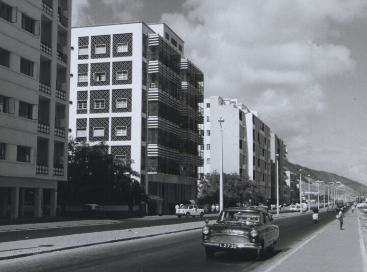 Neat multi-floored apartment blocks line a major road with cars driving by.