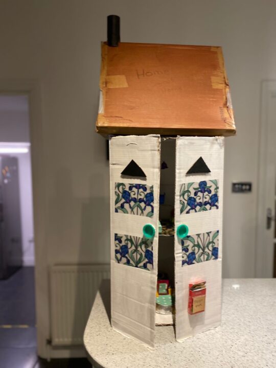 A completed model of the outside of the tall house made of cardboard. The roof is covered with brown paper and the walls decorated with designs to look like stained glass windows. A red letterbox is placed towards the bottom of the door.
