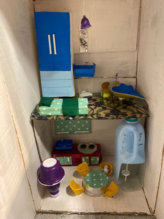 Inside of the two storeys of the model house. A lift in the corner is made from a plastic milk bottle, there are “kitchen units” and the upstairs features a bed, wardrobe, bath and toilet. The whole has been made using cardboard, foil, plastic lids and sweet papers.