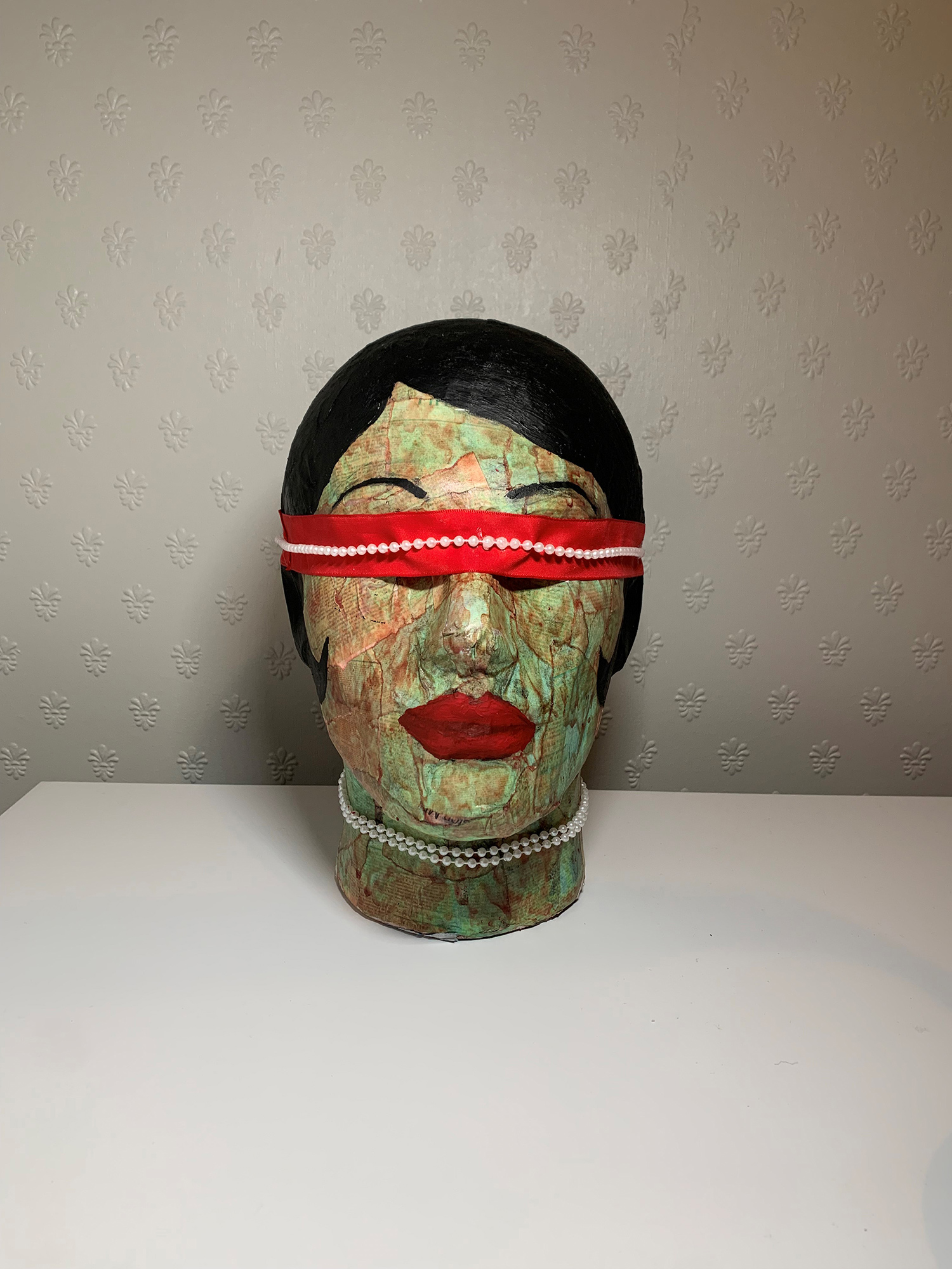Sculpture of a woman's head. She has short dark hair and red lipstick. She is wearing pearls around her neck and over her eyes.