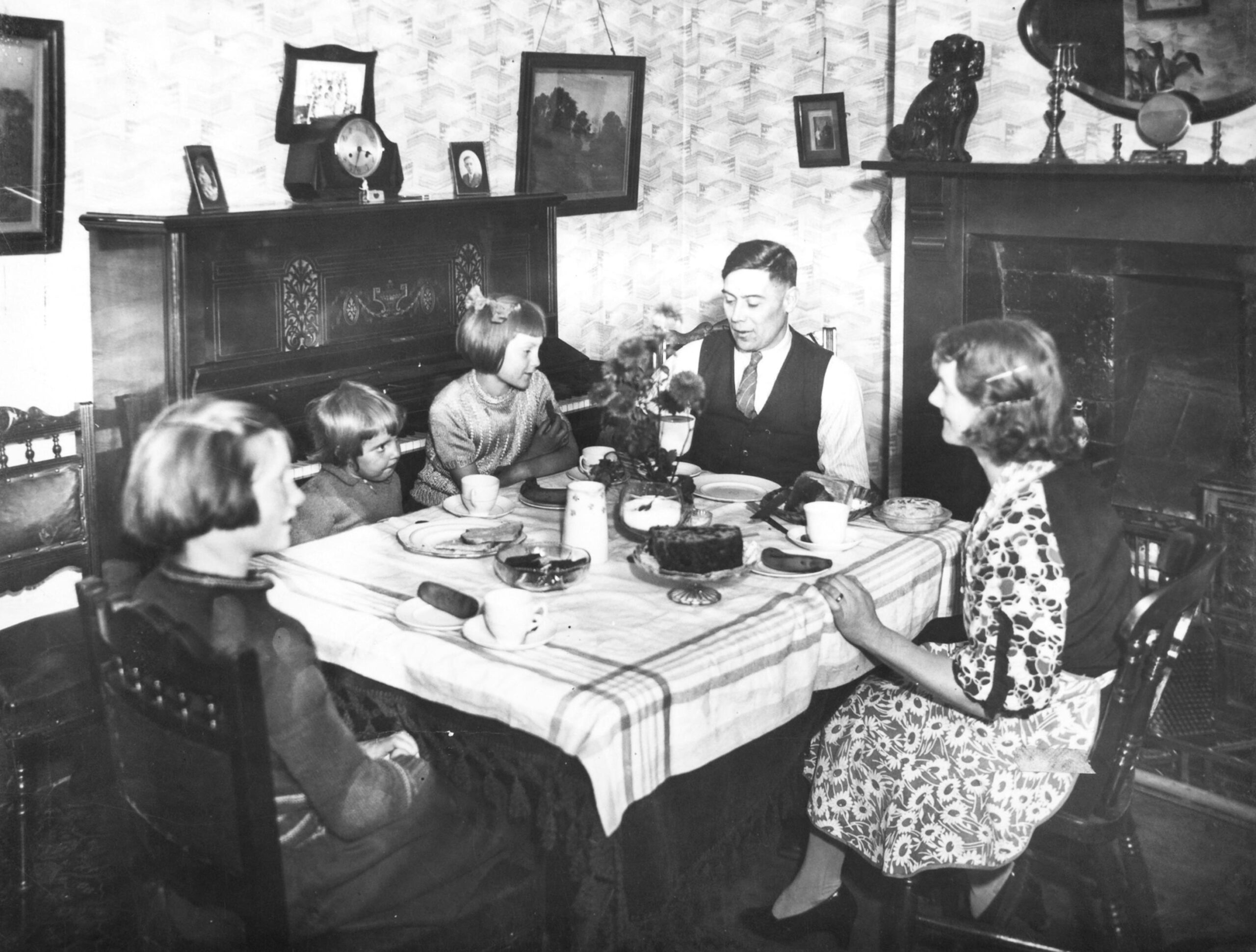A family consisting of two girls, one boy, and a mother and father sits around a table for a meal. They are white and dressed in nice clothes. The room is well decorated with a large fireplace and paintings on the walls.
