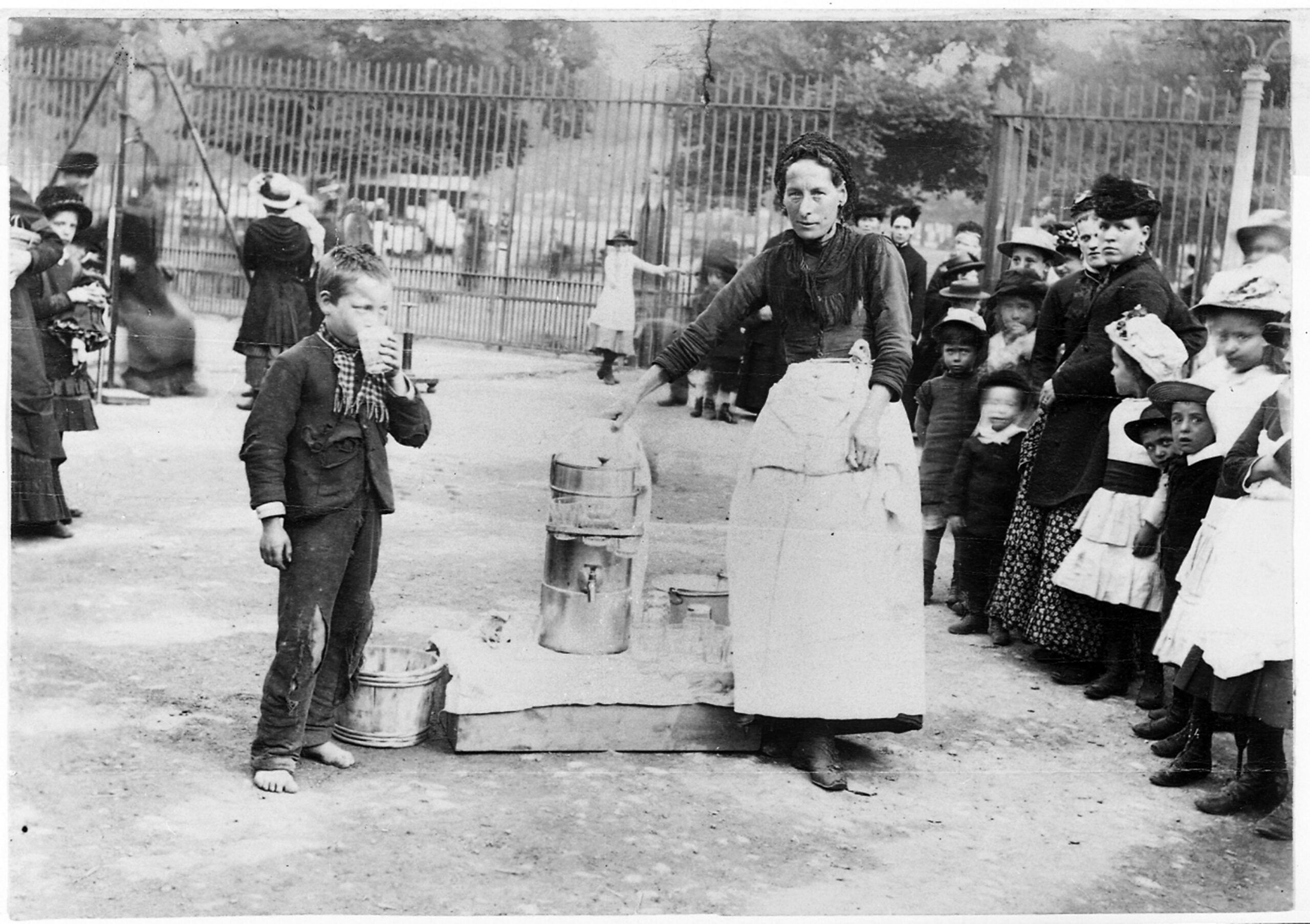 A woman stands by a large lemonade dispenser on the ground with glasses around it. A boy next to her drinks out of a glass. A crowd surrounds them.