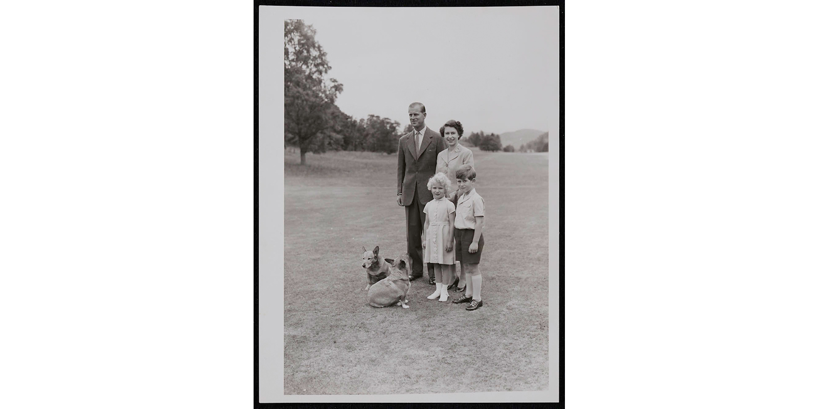 (left to right) Prince Philip and Queen Elizabeth II, with Princess Anne and Prince Charles standing in front of them. The four are posing for a photo in an open field. There are trees behind them in the distance, and two corgis at Philip’s feet. Elizabeth, Anne and Charles are looking at the camera, while Philip looks straight ahead.