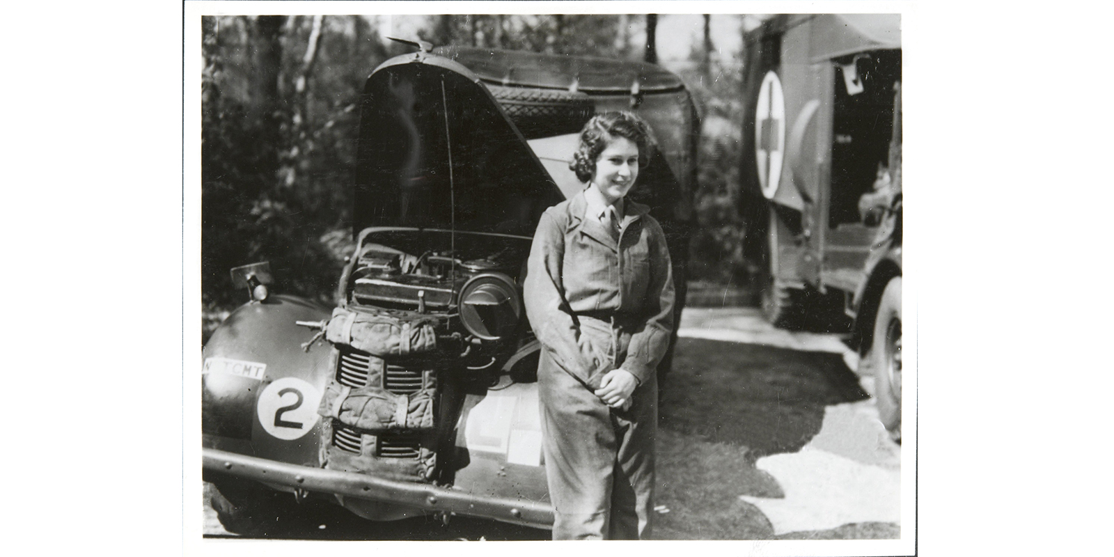 Princess Elizabeth leaning against the front wheel arch of a military vehicle, wearing a boiler suit. The bonnet of the vehicle is up, indicating that she has been maintaining it. A medical lorry is partially visible to the viewer’s right, adorned with a medical cross ‘+’ symbol on the side.