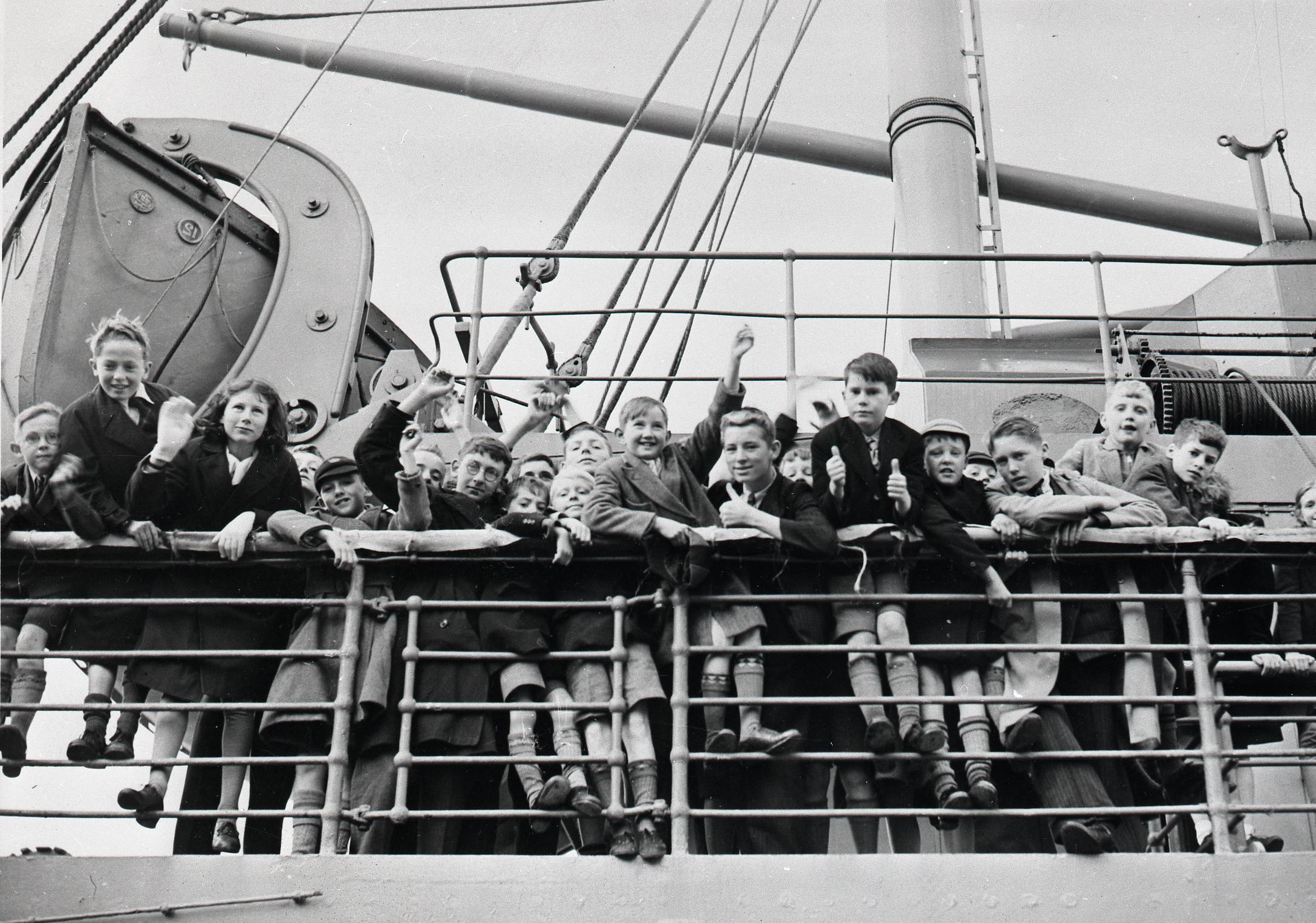 Monochrome photograph of a group of children waving and making thumbs up from the rail of a ship.