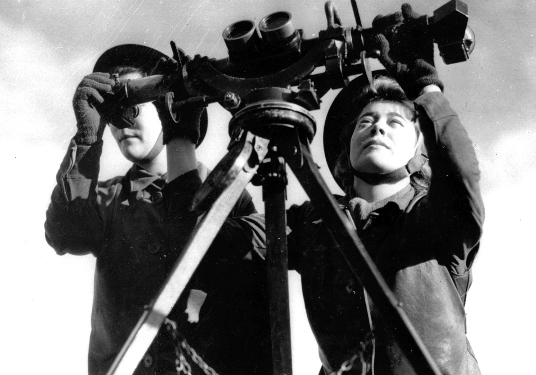 Monochrome photograph of two women in military helmets using an elaborate set of dual binoculars to look into the sky.