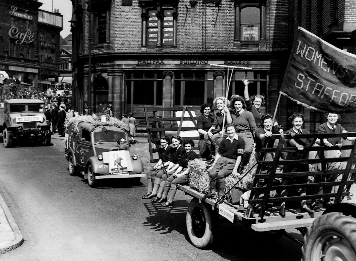 Monochrome photograph of women on a big tractor cart smiling and holding a banner that reads 'Women's Land Army Staffordshire'. Two cars follow behind them in a street lined with spectators. The car behind them has a large recruitment poster on its front.