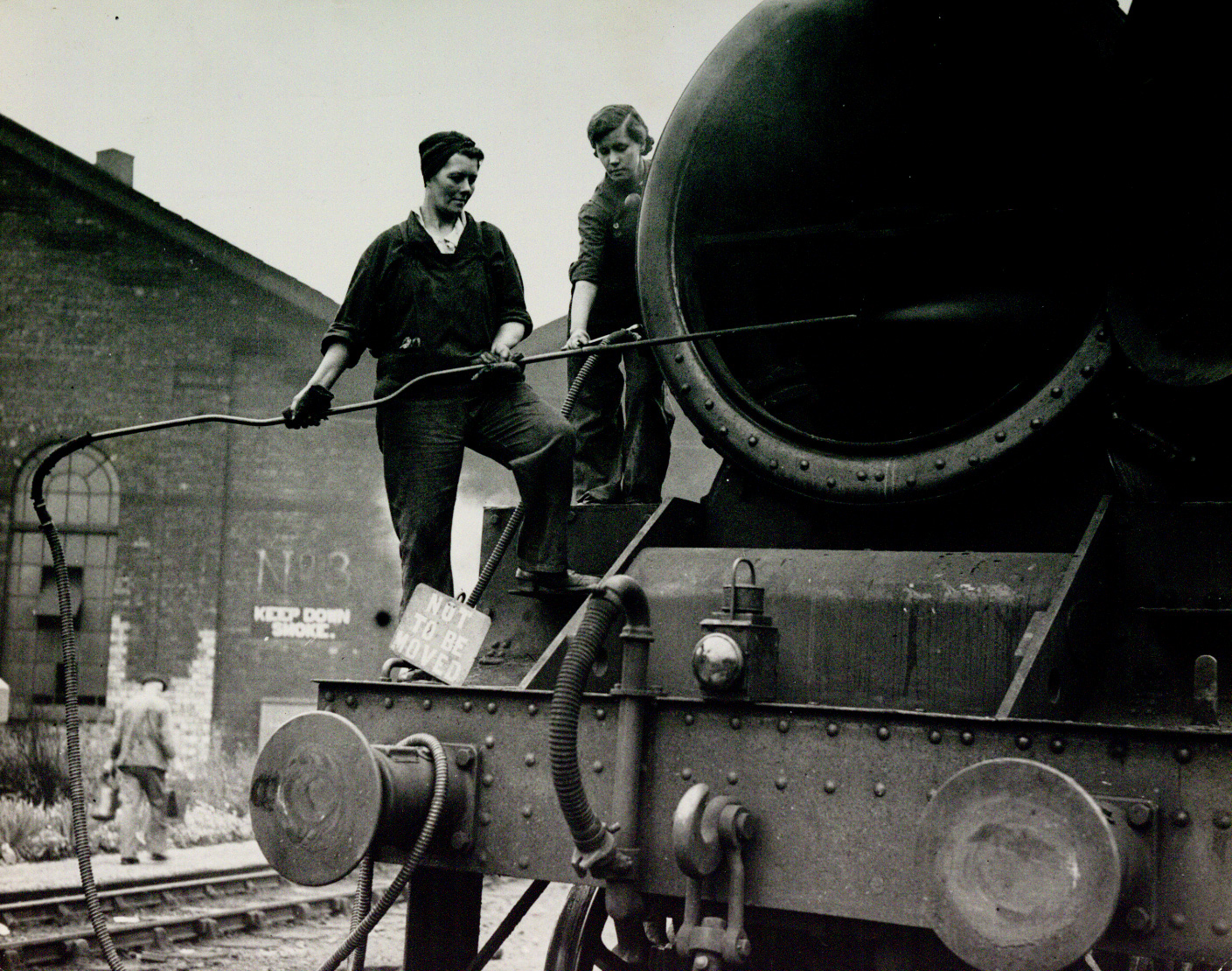 Monochrome photograph of two women standing on the wront of a steam locomotive and washing it with a pressure washer.