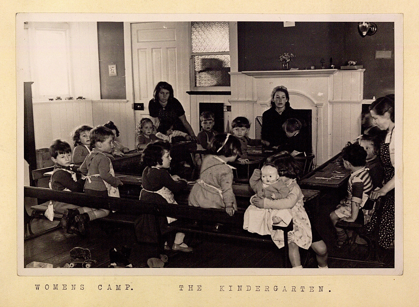 Monochrome photograph of a group of young children sitting at wooden benches inside a room.
