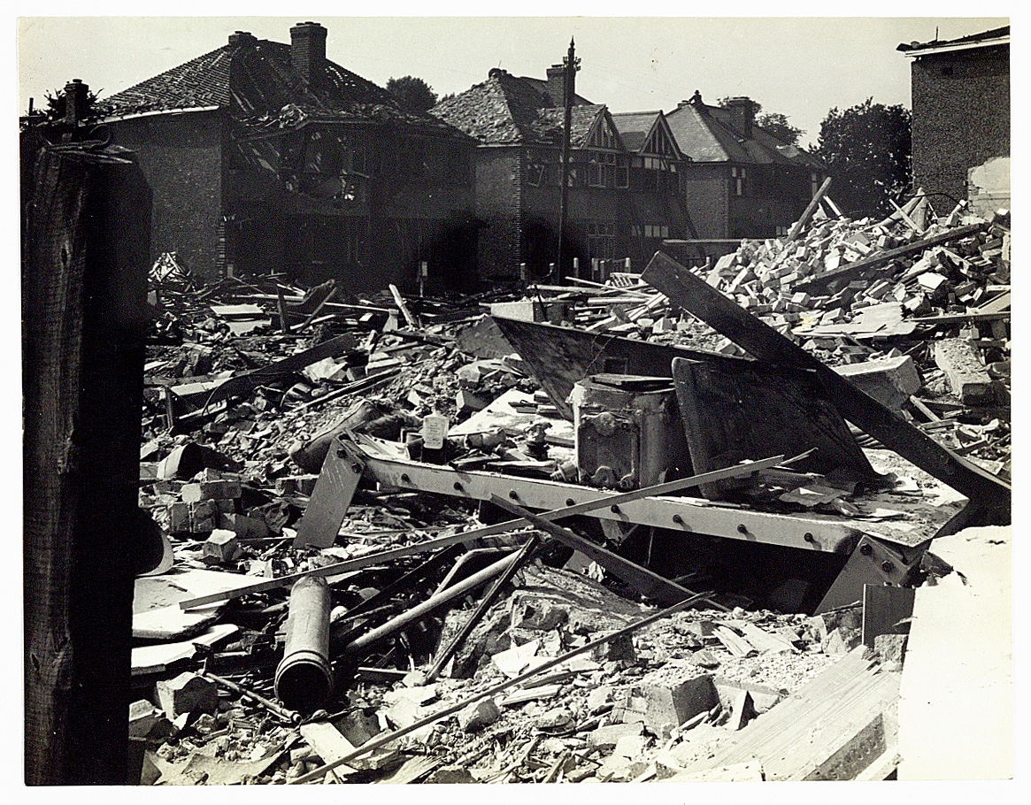 Monochrome photograph of a building reduced to rubble on a street with some houses that are still intact.