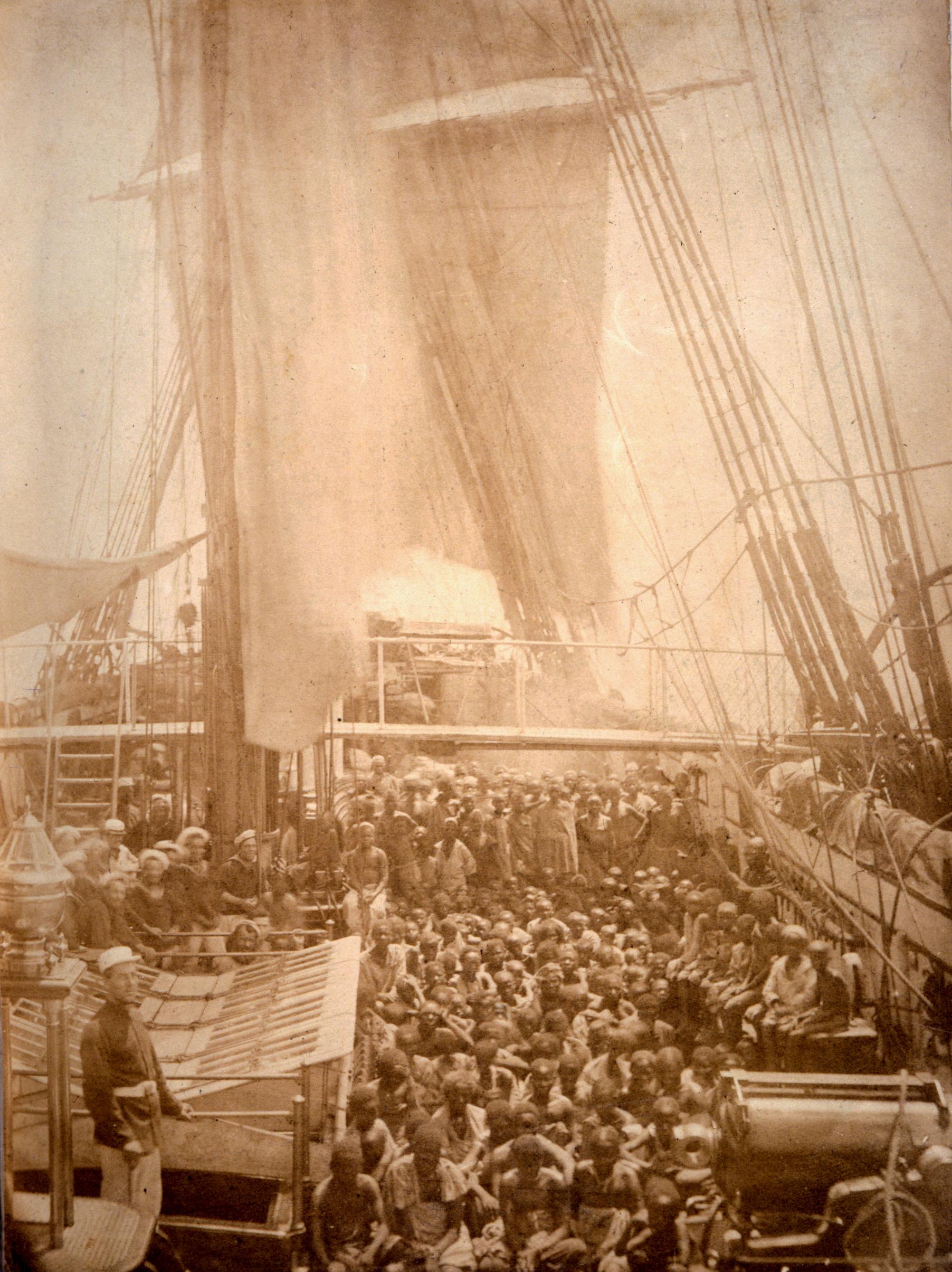 Sepia-toned photograph showing a large group of enslaved people, both adults and children, sitting and standing on the deck of a wooden ship facing the camera. A group of white sailors stand to the left.