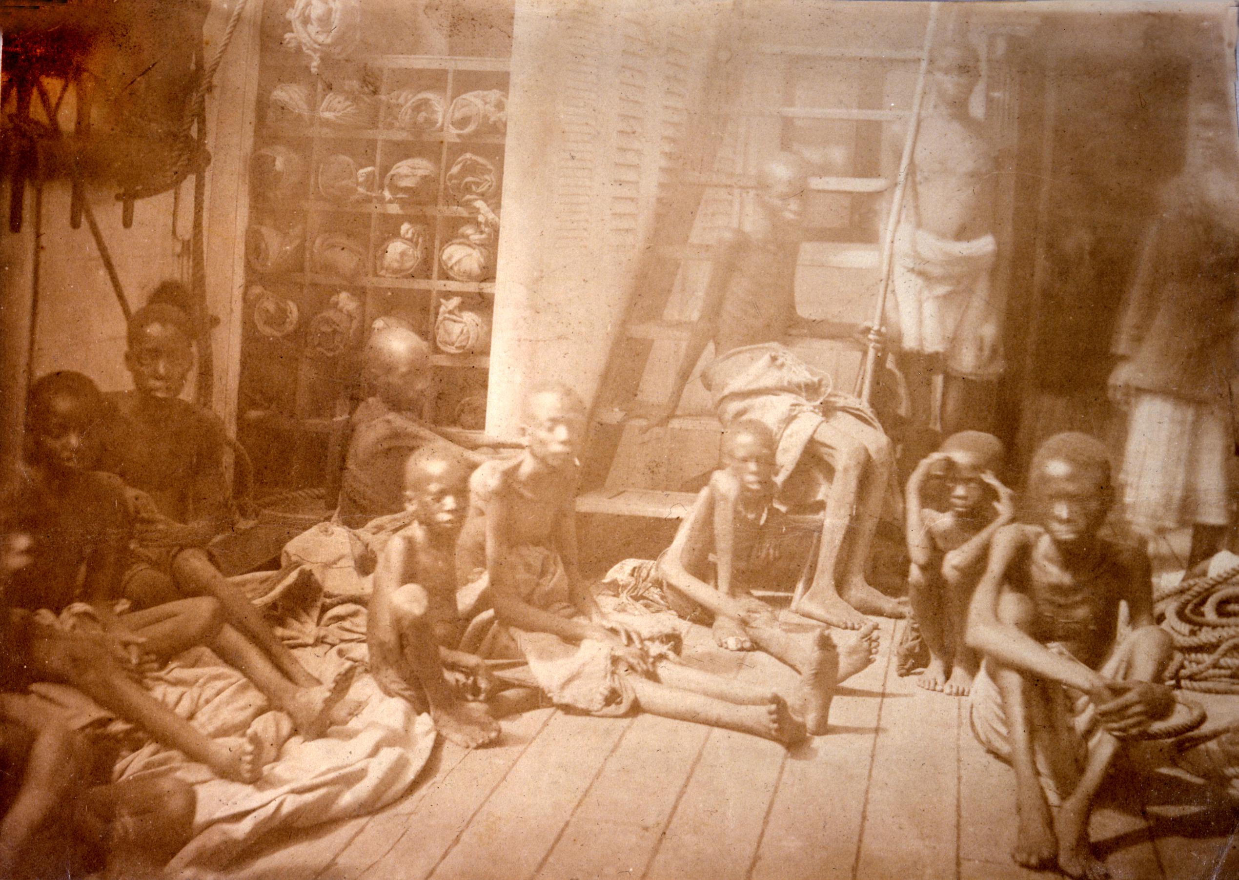 Sepia-toned photograph of a group of enslaved children sitting on the deck of a wooden ship. They are dressed in rags and appear emaciated.