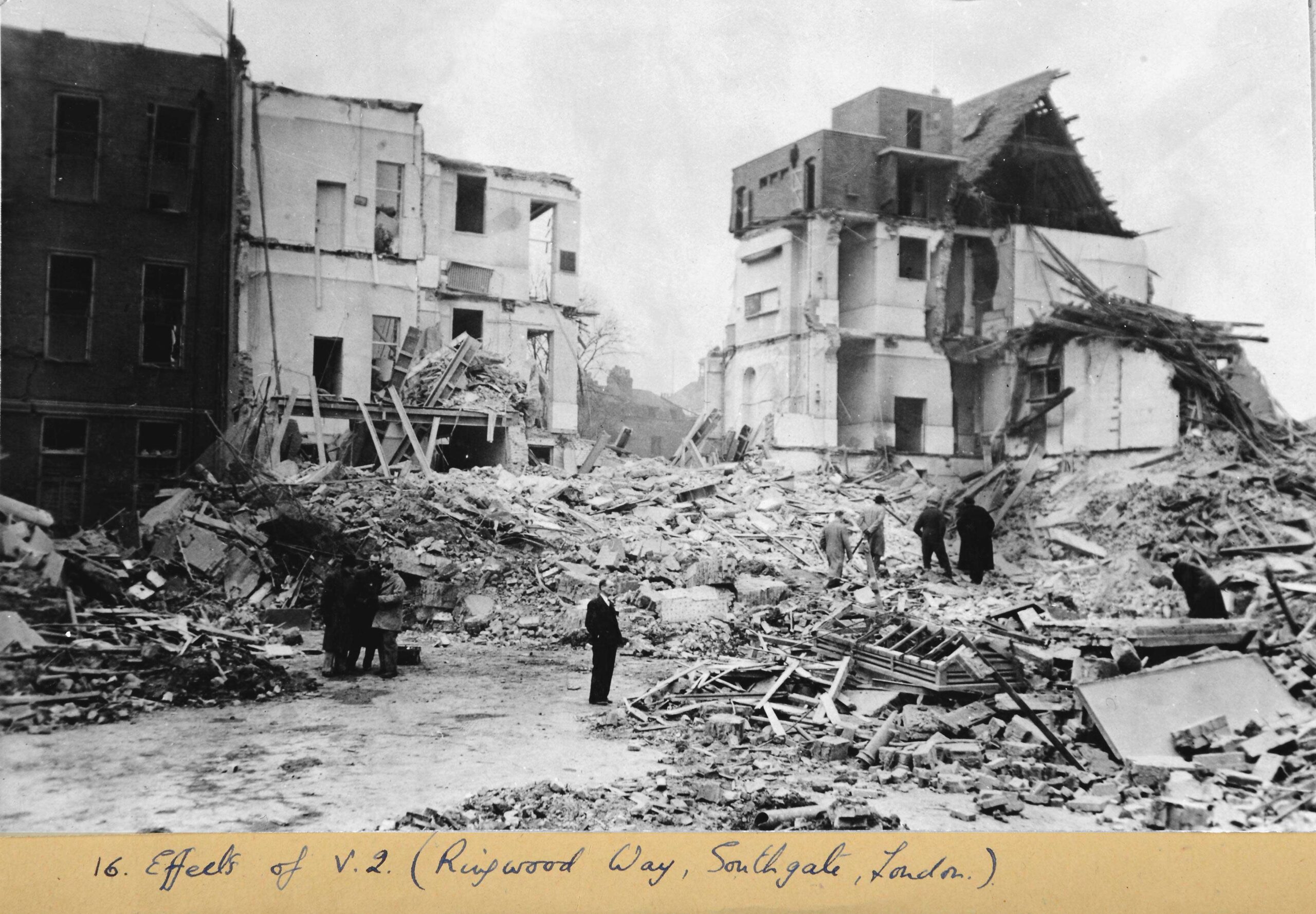 Monochrome photograph of a suited man standing in the middle of a city street reduced to rubble. Only a few houses are somewhat intact. Two groups of people close by stand talking or picking through the rubble.