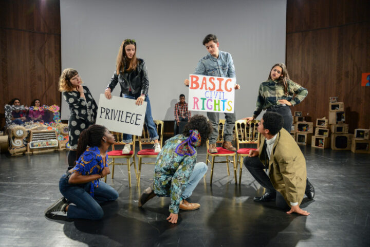 A group of people on a stage. Some are kneeling and some stand and hold signs saying "Privilege" and "Basic civil rights".