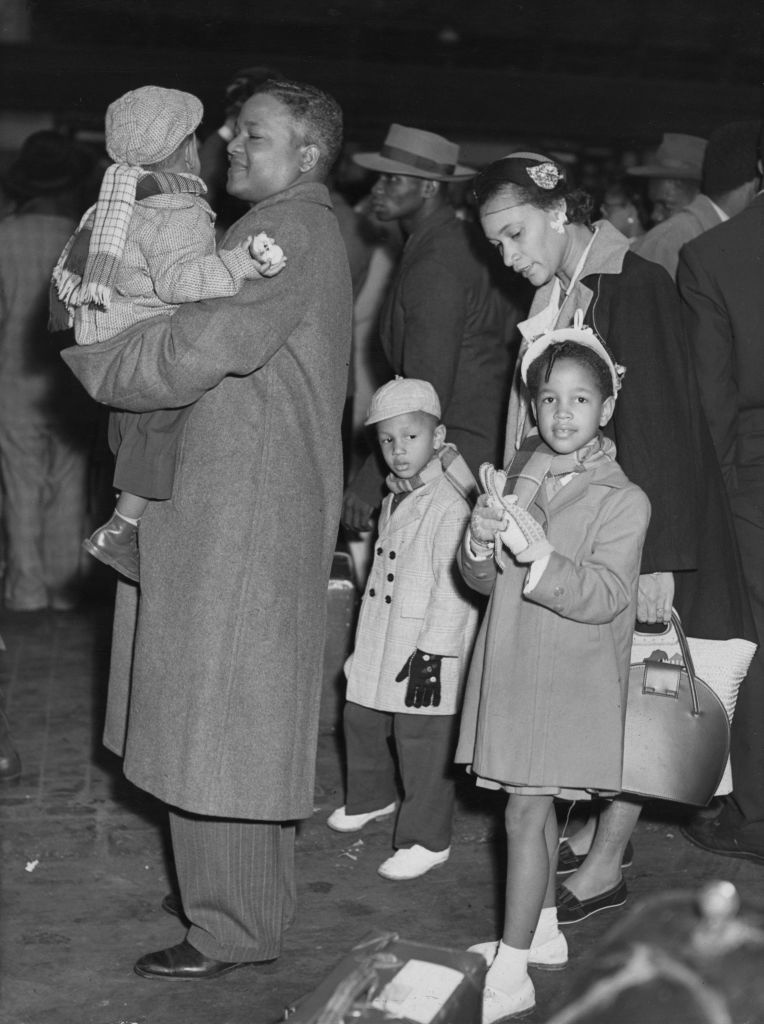 Monochrome photograph of a Black family who arrived in Britain in around 1950. The father is carrying a small child, there are two children with their mother walking just behind them. The family are dressed in winter coats, gloves and everyone apart from the father is wearing a hat. There is a suitcase visible and many people in the background, mostly wearing suits and hats. 