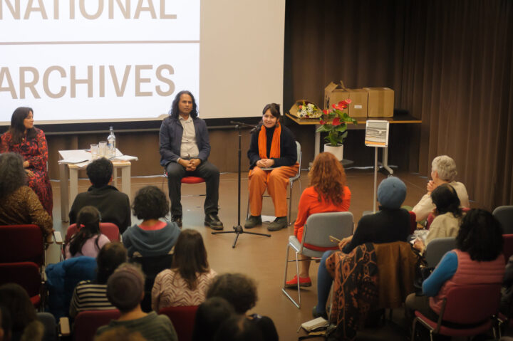 Two people sit on chairs in front of a crowd speaking into a microphone.