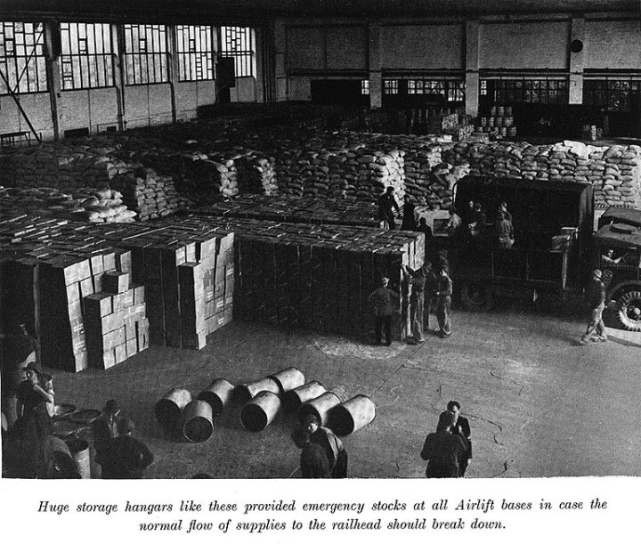 Interior photograph of a storage room or hangar with high windows with bars. The space contains boxes and filled sacks piled high. There are nine drums in the foreground. Three men stand nearby. An army vehicle is also being unloaded by two men in the storage hangar.