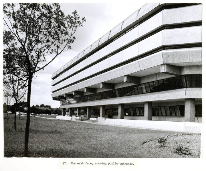 A large concrete building with two levels of larger windows and three levels with very small windows and concrete walls. Captions says 'The east face, showing public entrance.'