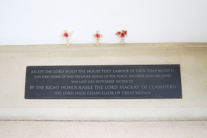 A metal sign on stone inside a building. Three crosses with poppies on have been placed on top of the sign.