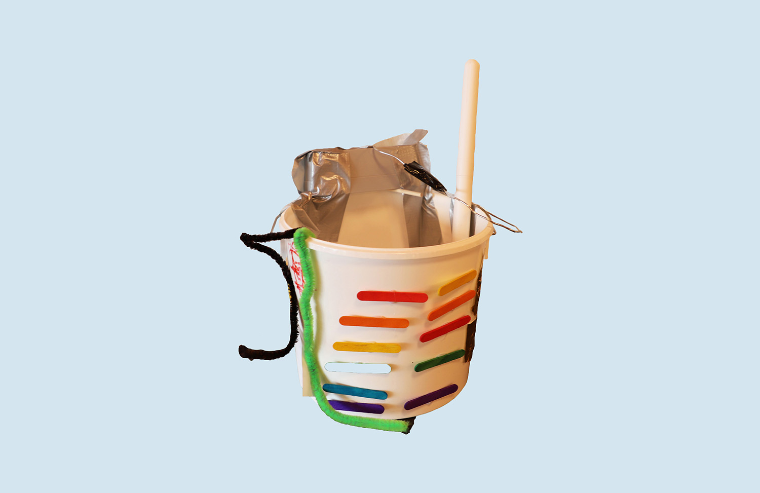 White plastic bucket with colourful ice cream sticks stuck on the outside. Two pipe cleaners stick out from the inside. The handle has been duct taped. A scrubber with a white handle has been placed inside.