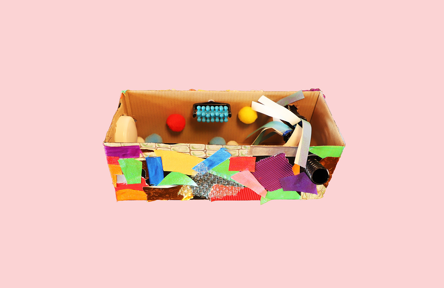 A cardboard box that has been opened to create a room. The room contains several sensory items including a green rubber mat, a blue knobbly wall feature, and colourful pompom balls. Lots of colourful paper is stuck on the outside walls.