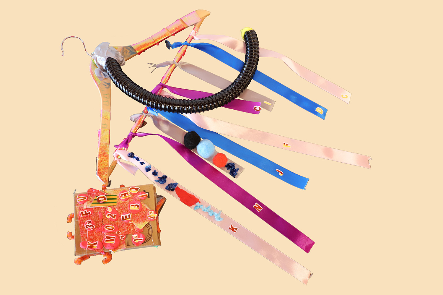 Hanger with multiple colourful ribbons attached, as well as a black tube. Next to it is as book-like object made out of cardboard with letters stuck on the front and orange pipe cleaners sticking out of it.