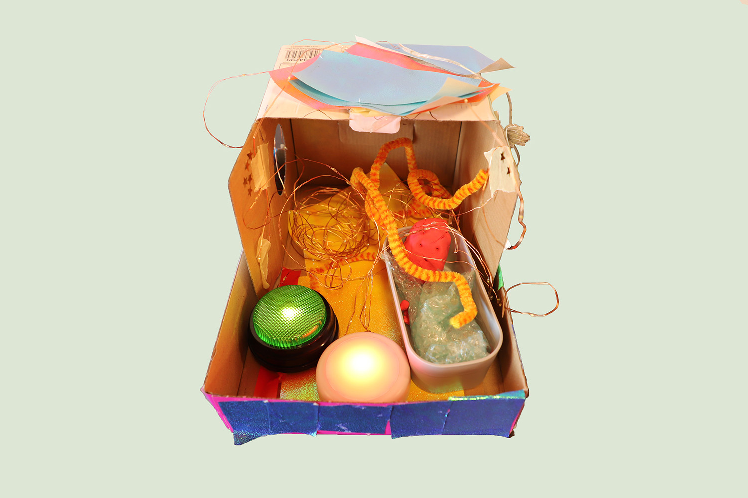 A cardboard box that has been opened to create a small room. It contains a small doll lying in a bathtub, a roll of fairy lights, and two large coloured lights. The floor and roof are lined with colourful paper.