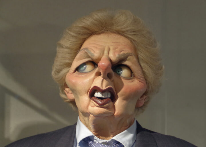 Head and shoulders photograph of ‘Spitting Image’ puppet created to represent Prime Minister Margaret Thatcher. She is wearing a dark suit, white shirt, and light blue spotted tie. Her face is flushed pink, her nose pointed, and her large blue eyes look wild. She is frowning. Her hair is light blonde.