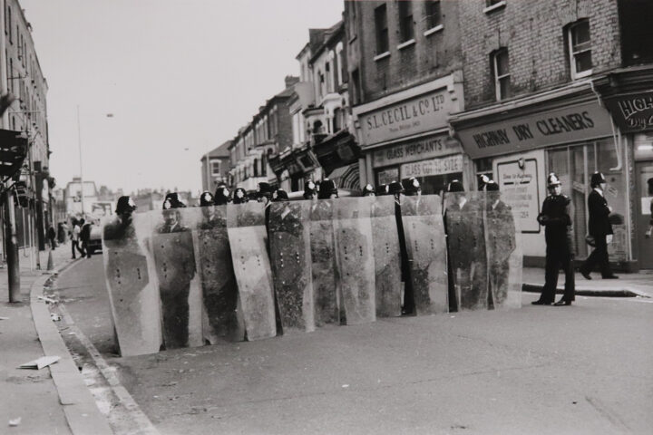 Monochrome photograph of a line of policemen holding riot shields in a street in Brixton.