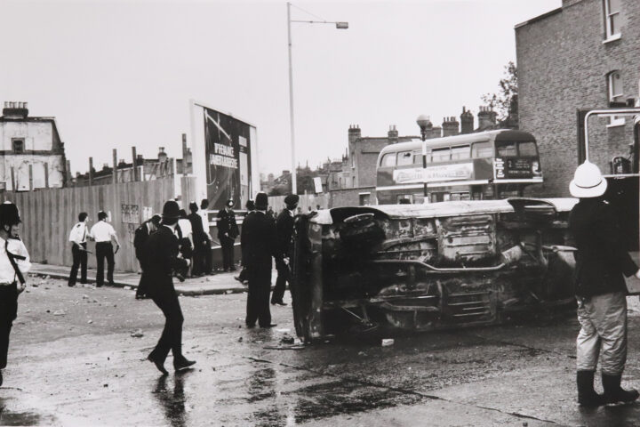 Monochrome photograph of policemen next to an overturned car on a street in Brixton.