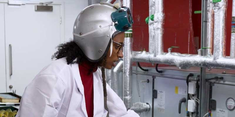 A woman wearing goggles and a silver helmet points at some equipment.