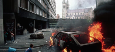 Young children play beside a burning car in the foreground with St Peter's Cathedral, Belfast, in the background. 