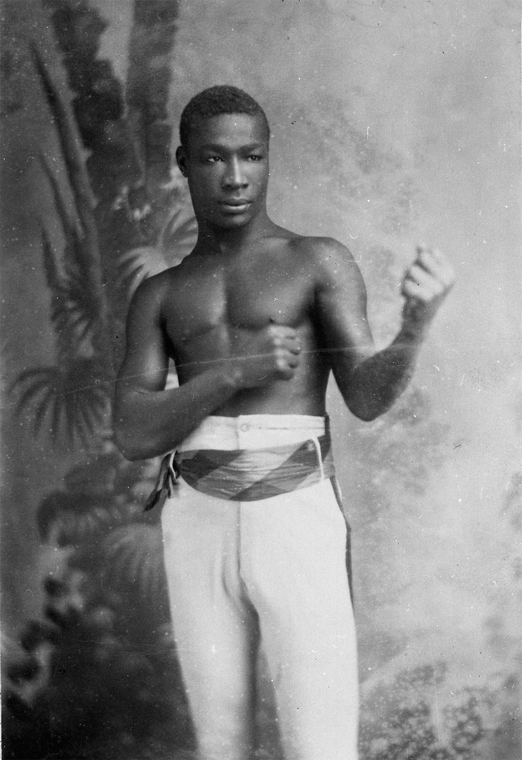 Monochrome photograph of a shirtless man wearing white trousers and holding his fists up in a boxing stance.