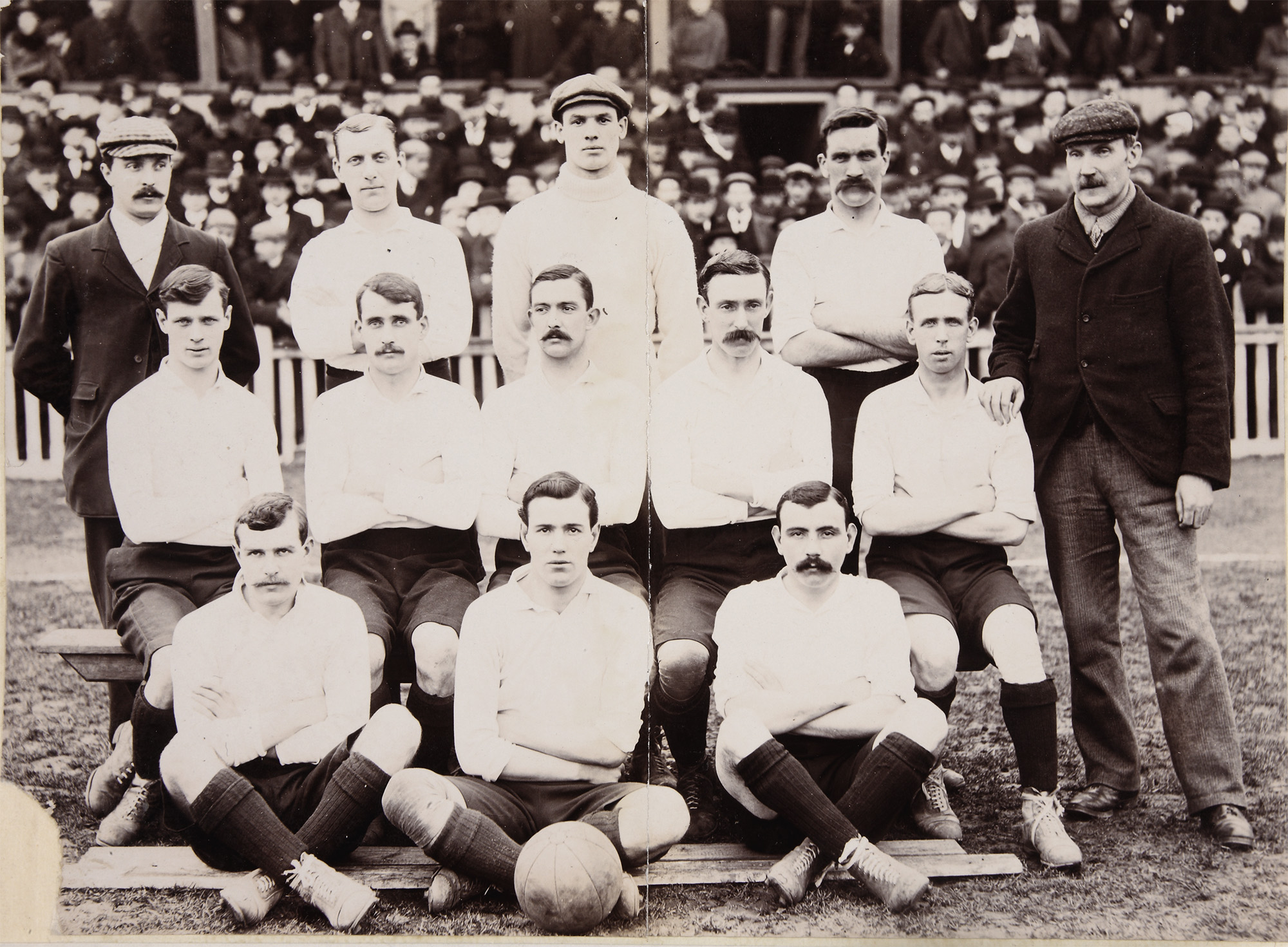 Monochrome group photograph of a football team. There are eleven players in uniforms of white shirt, shorts and knee socks and two other men in regular outerwear. A football lies on the ground in front of the team.