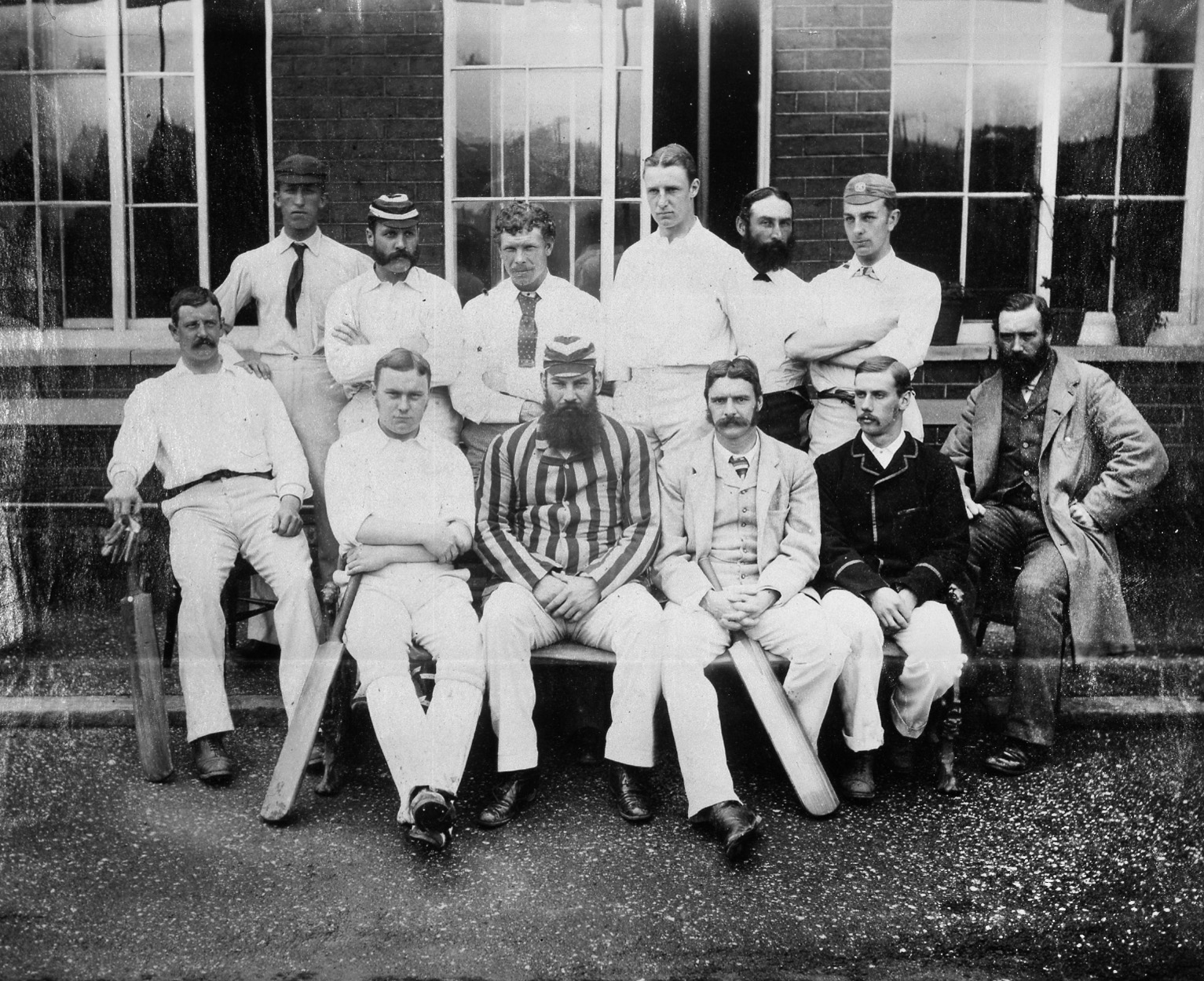 Monochrome group photograph of a cricket team consisting of twelve men. One is holding a cricket bat.