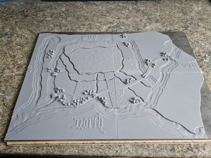 Tactile version of the map described above rendered in grey resin. The shape of the animal hide is copied. The houses on the map are rendered as small model villages along with small model bridges. The fields are rendered with a grass-like texture.
