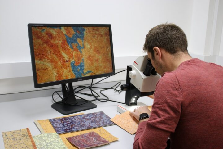 A male scientist looking through a microscope at orange and blue marbled paper. A close up image of the paper is shown on a computer screen.
