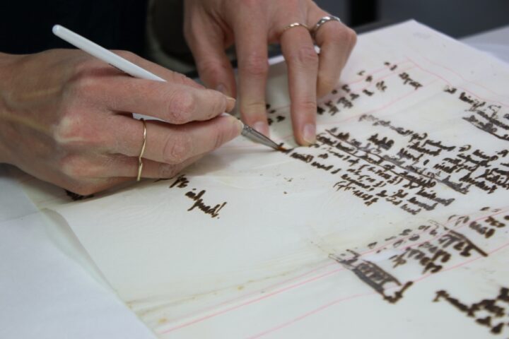 A conservator's hands are holding a very small paintbrush over the text on a paper document. They are conserving this text to ensure a clear image when the document is digitised.