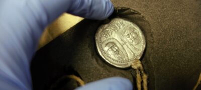 A blue-gloved hand reaches out to touch a metal seal encased in foam and depicting the faces of two men.