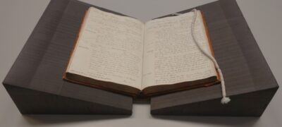 A book open on a table, supported by two triangular foam wedges and with a white snake weight curled over the edge of the right hand page.