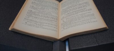 A paperback volume open on a table. It is supported by two triangular foam wedges. Between these wedges, a long black triangular spine insert supports the curve of the spine.