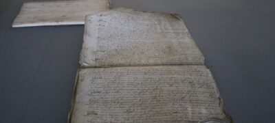 A folded parchment bundle on a table. One membrane has been unfolded flat onto the table, supported by a glass weight, and remains attached to the other folded membranes by its top left corner.